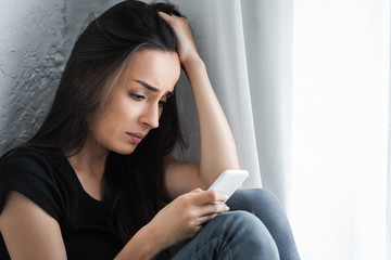 Obraz premium disappointed girl using smartphone while sitting by window at home and holding hand on head