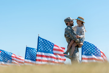 selective focus of man in military uniform holding in arms cheerful kid near american flags - obrazy, fototapety, plakaty