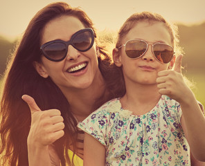 Happy fashion kid girl embracing her mother in trendy sunglasses showing thumb up and looking on nature background. Closeup portrait of happiness. Toned