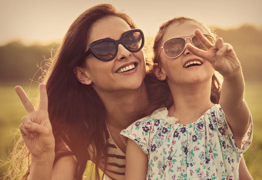 Happy fashion kid girl embracing her mother in trendy sunglasses showing victory sign and looking on nature background.. Closeup portrait