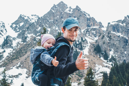 Attractive Young Father With Her Baby Daughter In Ergonomic Baby Carrier On Back Outside In Mountain Nature, Show Thumbs Up.