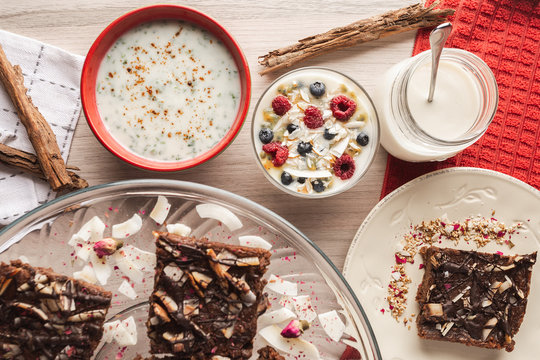 From above appetizing fragrant yogurt with raspberry currant oat in glass and red bowls decorated with cinnamon sticks and sliced brown cake on wooden background