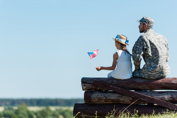 Patriotic child holding American flag near veteran father while sitting in fence