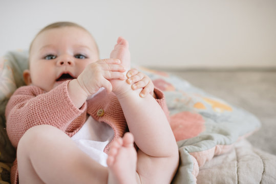 Charming cheeky baby in knitted blouse lying on blanket and catching small feet looking at camera in light room