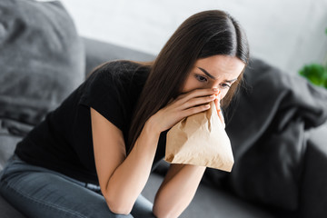 young woman breathing into paper bag while suffering from panic attack at home - obrazy, fototapety, plakaty