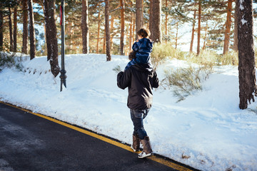 Side view of young man in dark clothes holding child in overall on neck and walking on yellow stripe on road along snowy forest