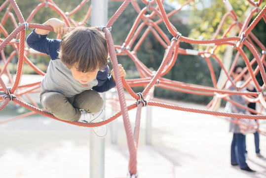 Pensive relaxed kid hanging on rope climbing net on playground in bright light