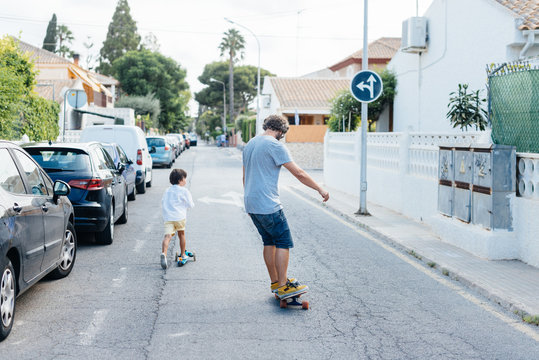 Back view of man on skateboard and boy on scooter riding down city street together
