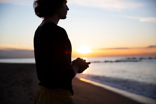 Side view of man concentrating on thoughts with closed eyes and hands in prayer gesture standing on knees on sandy beach in sunlight