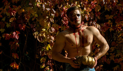Close-up portrait of a man with zombie makeup. Muscular man with broken head. Scary face man with horror Make up holding Pumpkin head jack lantern. The scary zombie is in the street with pumpkin.