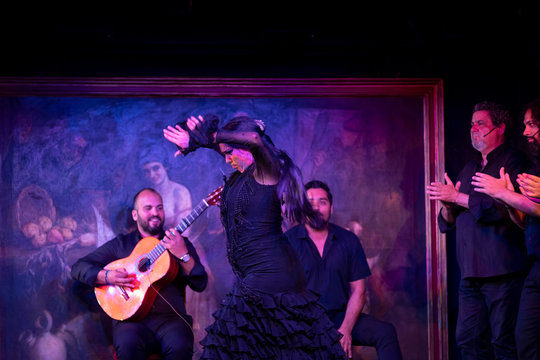 Woman in black costume dancing flamenco near Hispanic male musicians during performance against painting on dark stage