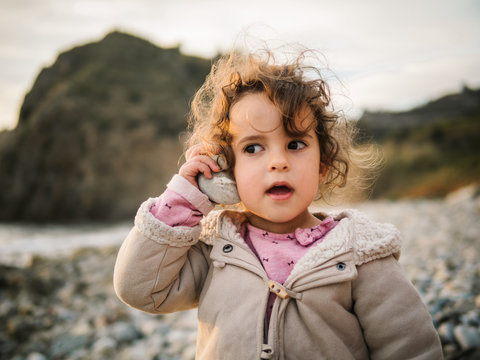 Portrait of cute female kid listening to conch with rapt attention while resting on stony seaside