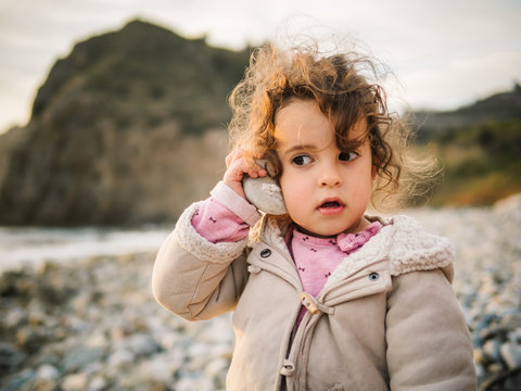 Portrait of cute female kid listening to conch with rapt attention while resting on stony seaside