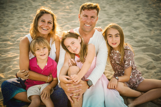 Adult loving man and woman with son and daughters sitting together on beach in back lit smiling at camera