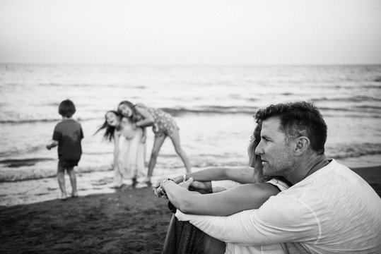 Adult loving man and woman sitting at the beach while their son and daughters play together next to sea, black and white photo