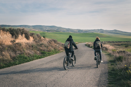 Back view of young men in dark clothes and backpack riding bicycles on empty road winding between stony hills in semi-desert Bardenas Reales Navarra Spain