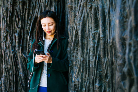 Beautiful Asian woman in stylish outfit listening to music and using smartphone while leaning on rough wall with relief of tree routs