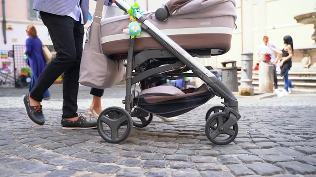Brown baby carriage with four wheels driving forward to the public park. Happy family walking slowlywith newborn child in the stroller on the cobblestone road. Modern comfortable baby pram 