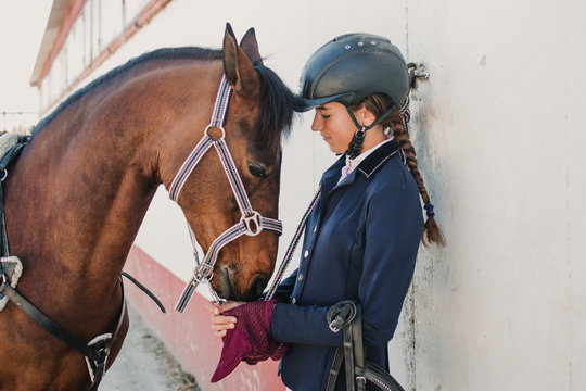 Side view of young teen woman in jockey helmet and jacket caressing horse standing together outdoors