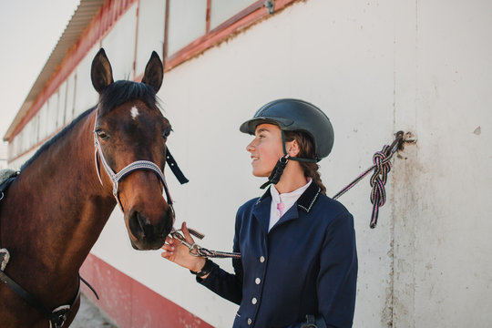 Side view of young teen woman in jockey helmet and jacket caressing horse standing together outdoors