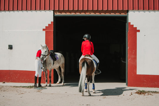 Back view of anonymous young teen woman in jockey helmet and jacket riding a horse standing next to stable entrance