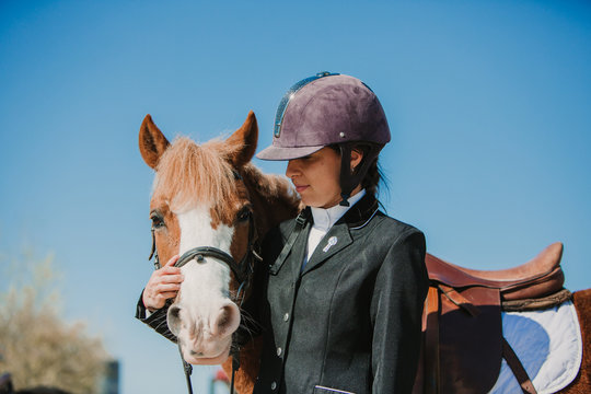 Side view of young teen woman in jockey helmet and jacket caressing horse standing together outdoors against blue sky