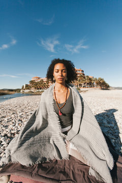 African American attractive young woman covered with grey plaid meditating in lotus yoga posture on sandy beach in bright day
