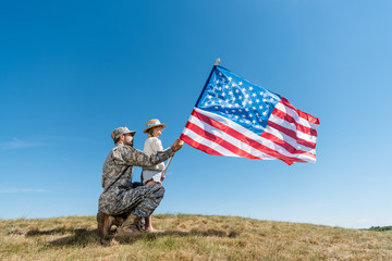 Father in military uniform and kid holding American flag against sky