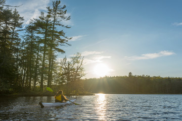 white kayak with a woman at the helm paddles to the sunset in an Adirondack lake