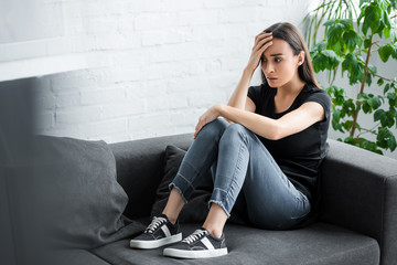 pretty young woman suffering from depression while sitting on couch at home and holding hand near...