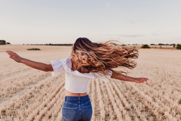 Summer Girl enjoying nature on yellow field. Beautiful young woman dancing Outdoors. Long hair in the wind. Happiness and lifestyle. Back view