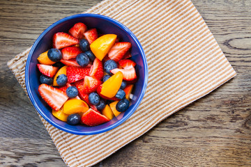 Fototapeta na wymiar Top down view of a colorful bowl of mixed fruit salad with bite sized pieces; Bowl of cut up fruit with a tea towel on a wood surface