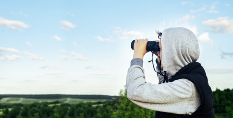 Obraz premium A man with modern binoculars against the sky and green hills. The concept of hunting, travel and outdoor recreation. Banner with copy space.