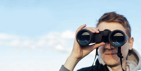 A man with modern binoculars against the sky and green hills. The concept of hunting, travel and outdoor recreation. Banner with copy space.