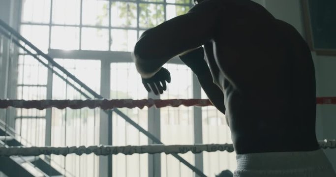 Cinematic slow motion shot of a professional young muscular shirtless african man is practicing shadow boxing work out in a gym with ring. 