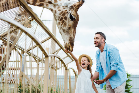 Selective focus of cheerful man and kid with closed eyes feeding giraffe in zoo