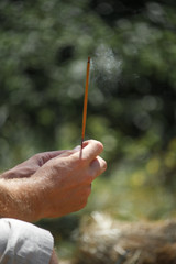 Hand of a man holding a burning incense stick on blurred natural background, selective focus