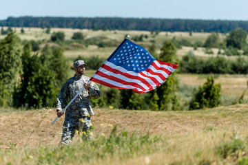 selective focus of handsome military man in uniform and cap holding american flag in summertime