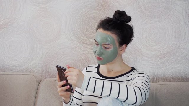 A young girl with a mask of white clay sits on the couch and uses a smartphone.