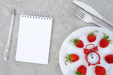 Red ripe strawberries berry on white plate, cutlery, red alarm clock and open blank notepad on gray stone table. Top view, flat lay, mockup. Concept diet and detox time or summer menu time.