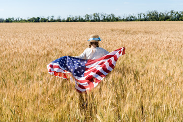 back view of kid holding american flag with stars and stripes in golden field
