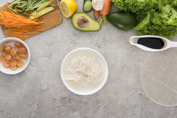 top view of bowl with water and noodles, lemon, avocados, carrots, black pepper, shrimps, soy sauce, rice paper, garlic, sliced vegetables and lettuce on table