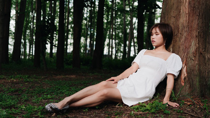 Portrait of beautiful Asian woman sitting under tree in summer forest, Chinese girl in white dress sleeping with eyes closed, relax time on holiday concept.