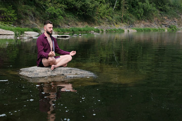 A young man of like Caucasian appearance in a plaid shirt is sitting on a stone in a river or lake. The concept of yoga, relaxation, unity with nature, travel and lifestyle. Copy space.