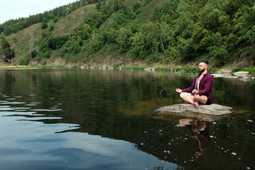 A guy with a kakvkazskoy appearance in a shirt with long sleeves sits on a stone in a river or lake. The concept of yoga in the forest, relaxation in the mountains, travel and lifestyle. Copy space..