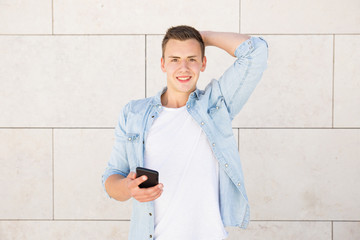 Portrait of cheerful young man standing at wall with cellphone. Caucasian guy using mobile phone outdoors. Mobile communication concept