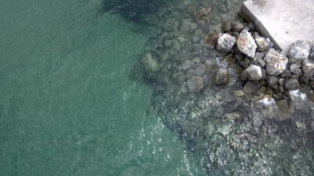 Aerial view of stunning turquoise sea with pier and rocks that water pops up on them in the area of Agia Paraskevi Halkidiki, Greece, up and side movement by drone