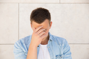 Tired young man touching forehead at wall outdoors. Handsome white guy wearing jeans shirt and keeping eyes closed with building wall in background. Stress or problem concept. Front view.