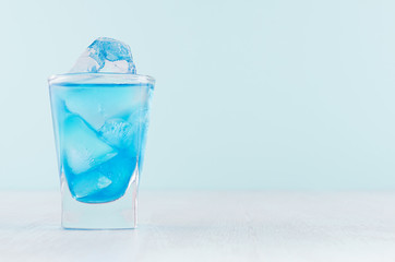 Refreshing alcohol drink blue Hawaii in misted rich shot glass with ice cubes in modern pastel mint beach bar interior.