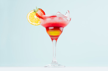 Summer layered drink with red and yellow liquor, orange slice, strawberry, ice cubes in wineglass on pastel mint background.